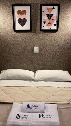 A hotel room with a bed, two pillows, a quilted mattress, and a set of towels with the hotel logo. There are two framed artworks on the wall above the bed. - Living Hotel São Paulo (São Paulo)