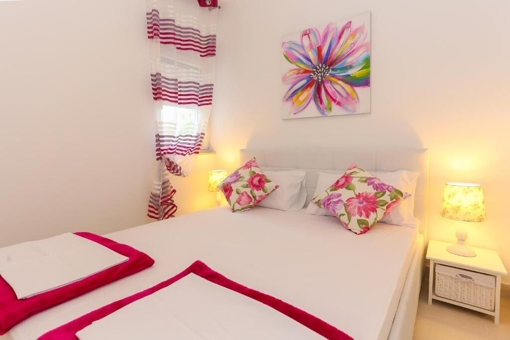 Quarto com cama, almofadas, mesa de cabeceira, abajur, janela com cortinas e um quadro com flores na parede.
