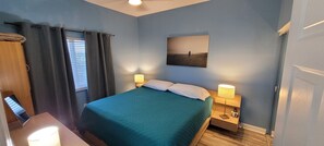 Une chambre à coucher comprenant un lit, deux lampes de chevet, une table de chevet, une fenêtre avec des rideaux et un tableau accroché au mur.