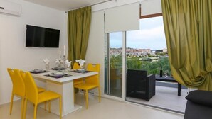 Ein moderner Essbereich mit gelben Stühlen und einem weißen Tisch, einem Flachbildfernseher an der Wand sowie ein Balkon mit Blick auf eine Stadtlandschaft.