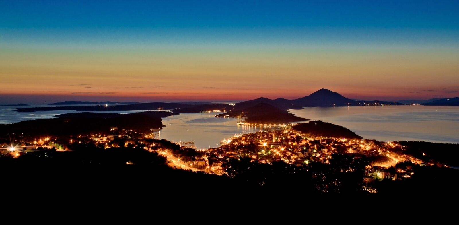 Un pueblo costero al anochecer, con edificios iluminados y una montaña en el fondo.