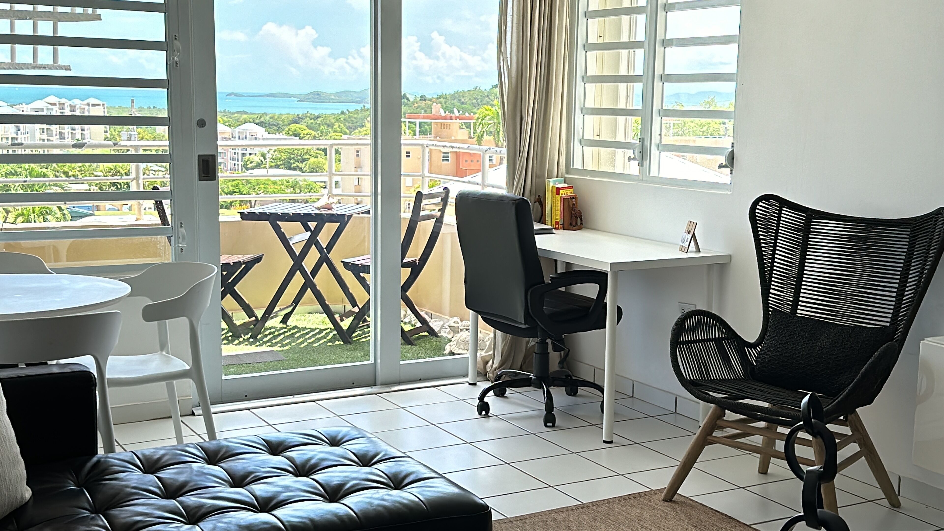 A room with a balcony, a dining table, a black leather ottoman, a black office chair, and a wicker chair.