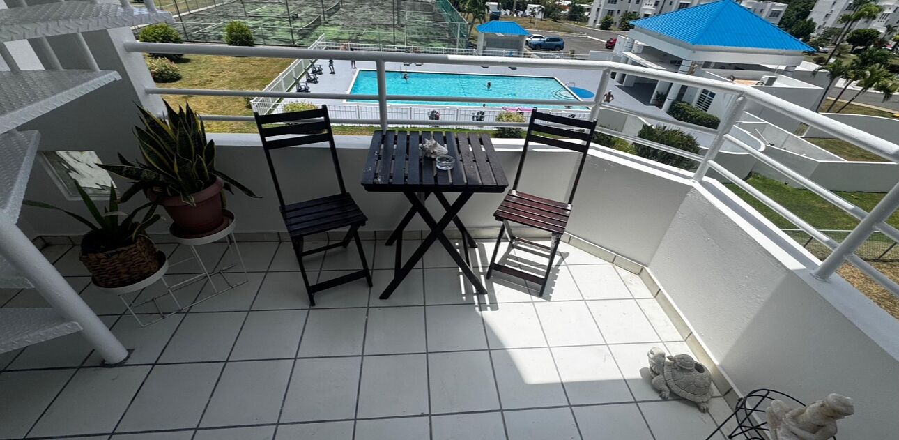A balcony with a table and chairs, potted plants, and a view of a swimming pool and buildings.