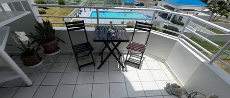 A balcony with a table and chairs, potted plants, and a view of a swimming pool and buildings.