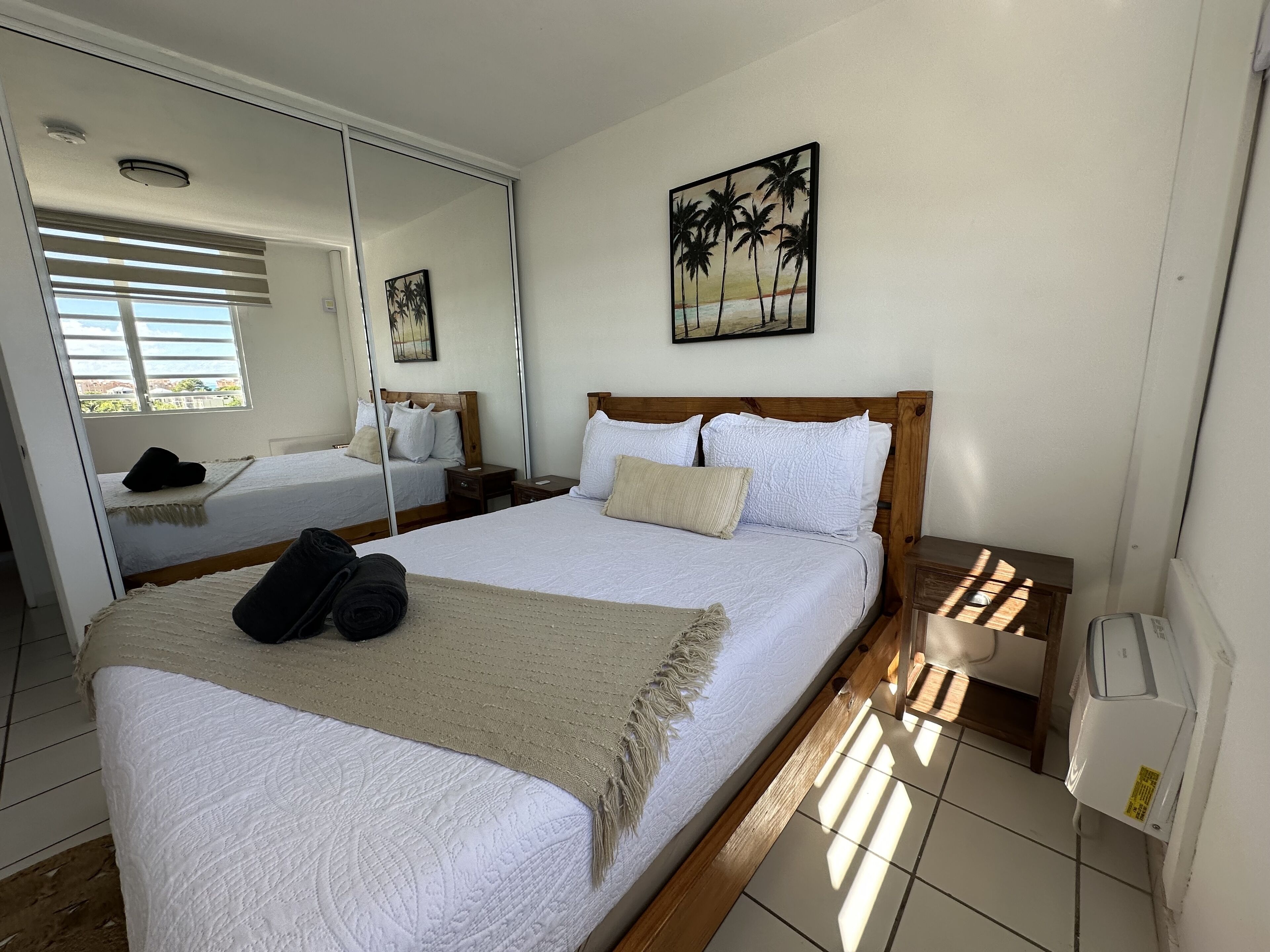 A hotel room with a large bed, a wooden headboard, a bedside table, a nightstand, a mirror, and a painting of palm trees.