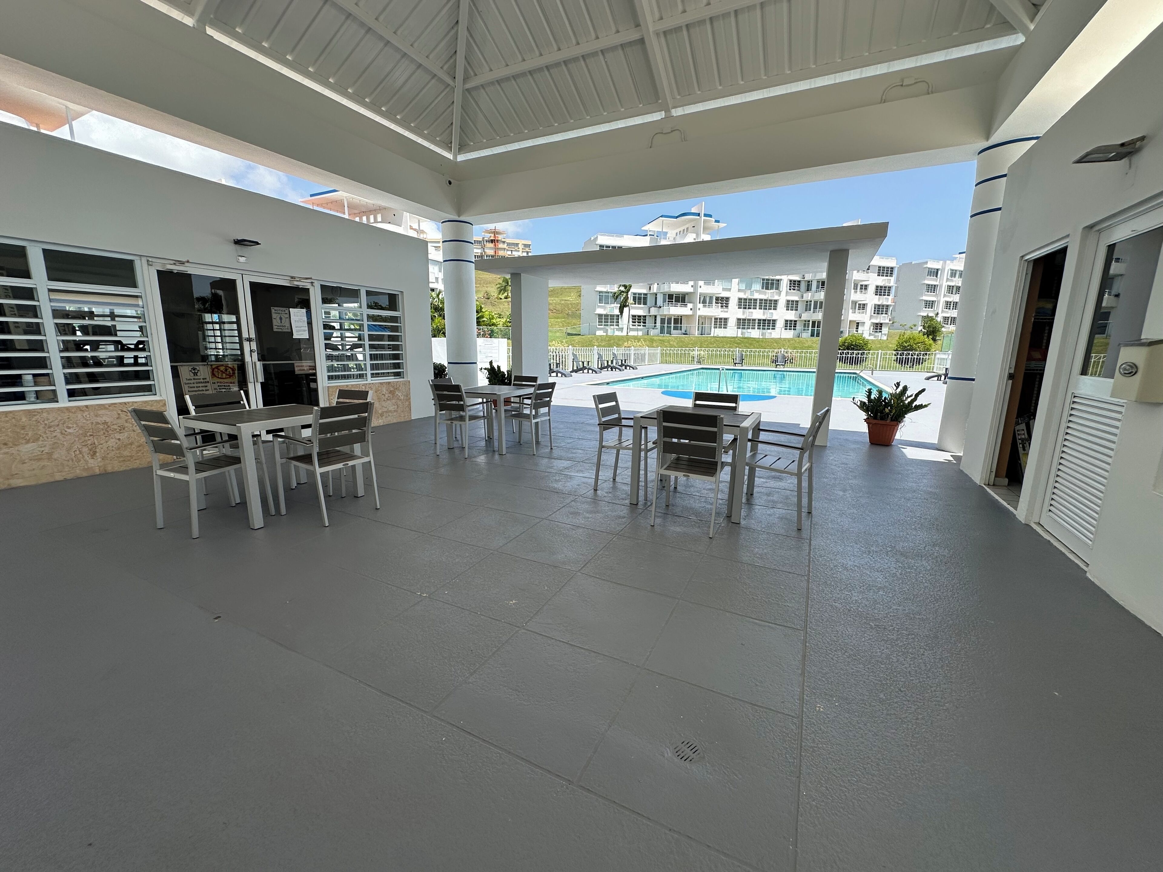 Un espace extérieur abrité, doté de tables et de chaises, avec une piscine en arrière-plan et une vue sur des immeubles d’habitation.