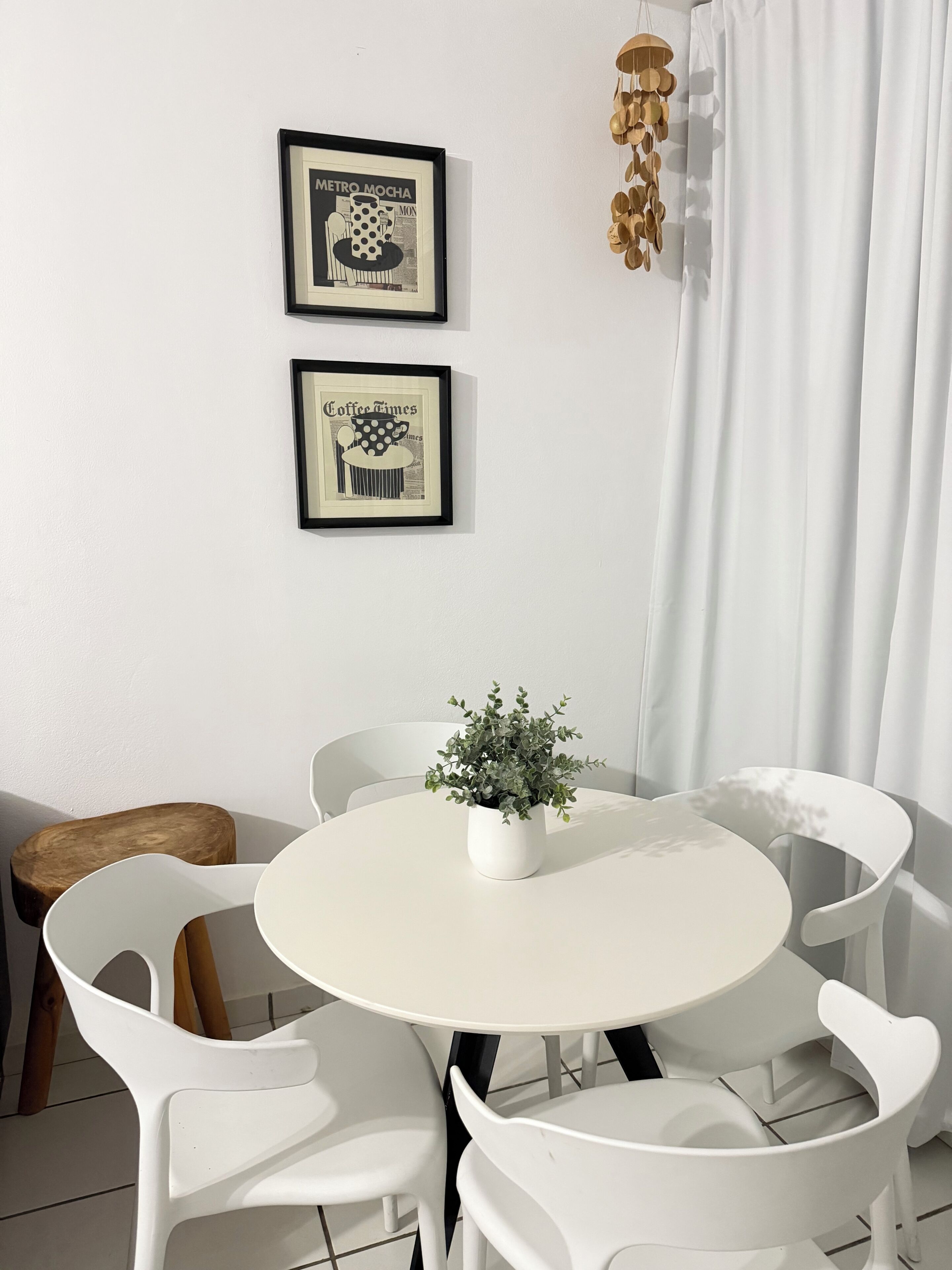 Een ronde witte tafel met een bloempot, omringd door witte stoelen, voor een witte muur met twee afbeeldingen in lijsten.