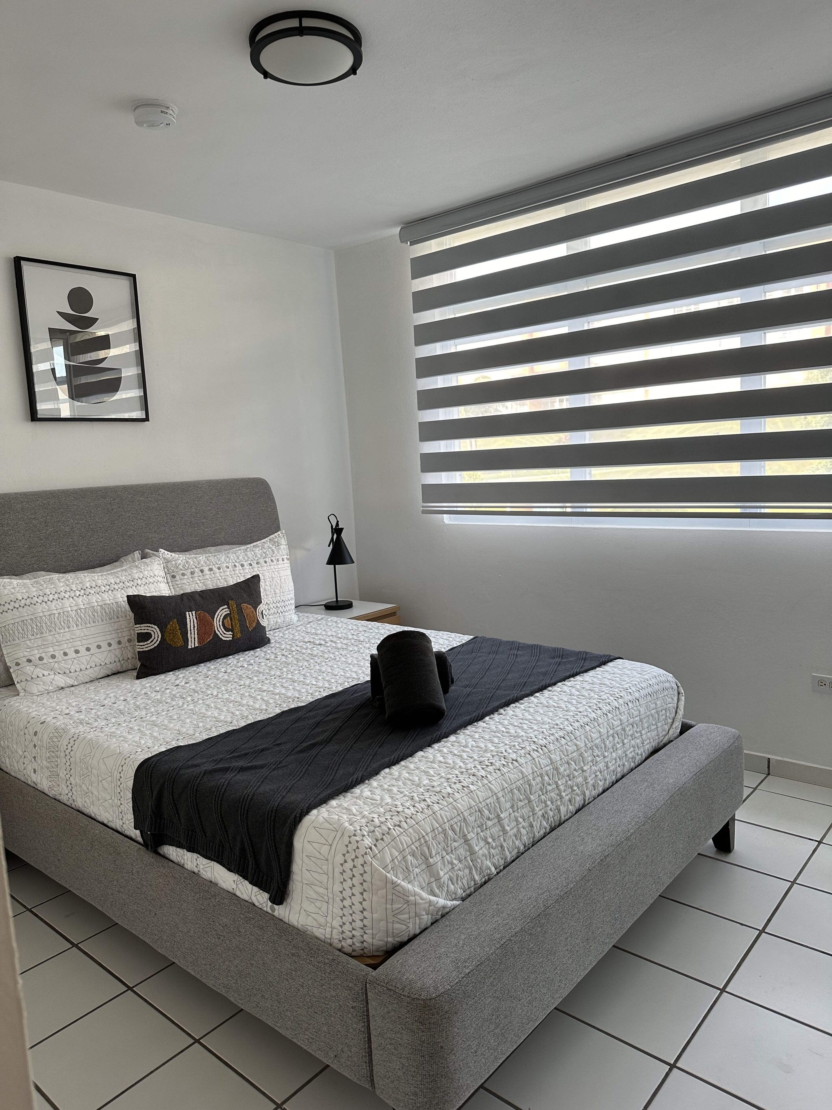 Une chambre moderne avec un grand lit, une œuvre d’art abstraite encadrée, une fenêtre avec des stores et un luminaire au plafond.