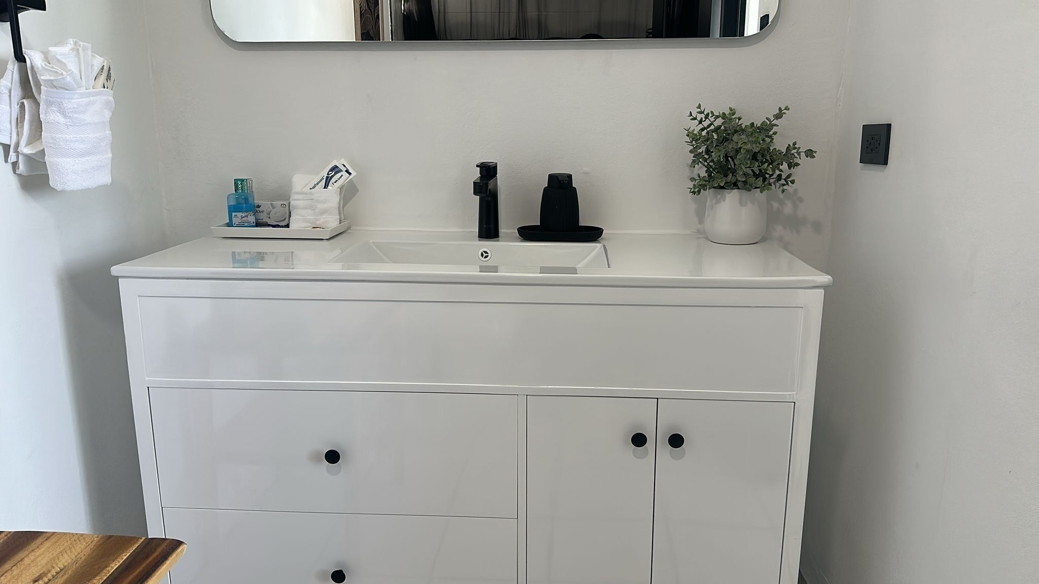 Une commode blanche avec un lavabo, deux robinets noirs, une petite plante et un porte-mouchoirs contenant des mouchoirs.