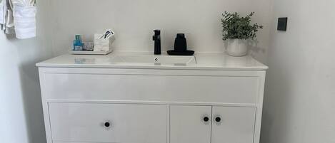 Une commode blanche avec un lavabo, deux robinets noirs, une petite plante et un porte-mouchoirs contenant des mouchoirs.