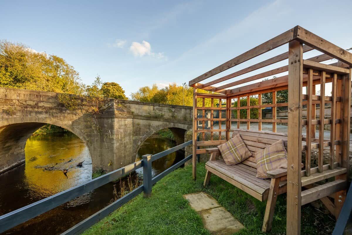 A wooden bench with a metal railing by a river, under a bridge.