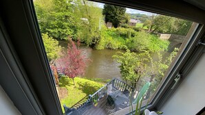 Blick aus einem Fenster auf einen Fluss, ein Holzdeck und einen kleinen Garten mit einem Baum in roter Farbe.