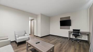 Una habitación moderna con suelo de madera, un sillón blanco, una mesita con un mando a distancia, un escritorio con una silla y un televisor de pantalla plana montado en la pared.