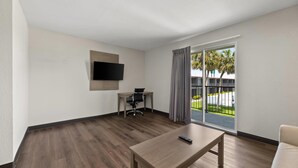 A modern hotel room with a wooden floor, a flat-screen TV mounted on the wall, a desk with a chair, a large sliding glass door to the outside, and a remote control on a wooden table. - Quality Suites Wildwood - The Villages (Wildwood)