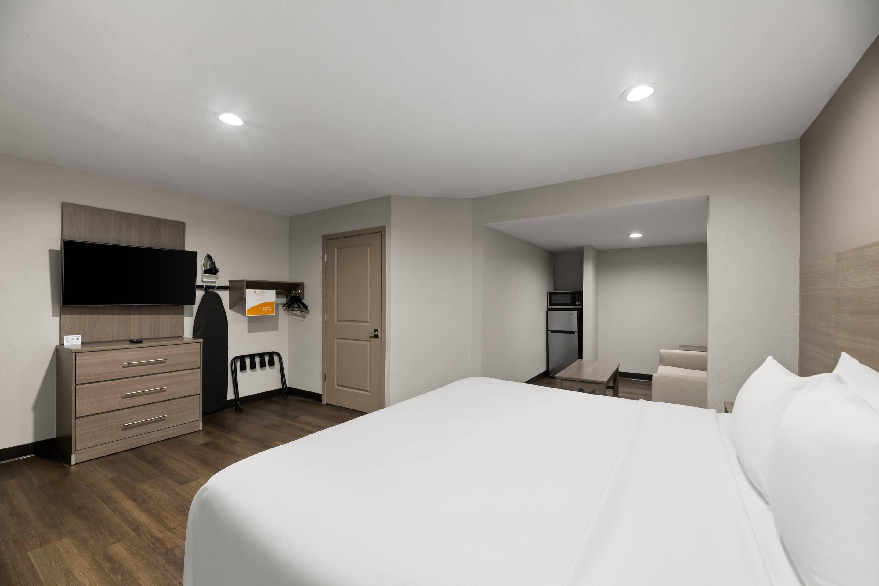 A modern hotel room with a large bed, a TV, a wardrobe, a desk, and a bathroom visible through an open door.