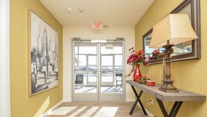 A hallway with a yellow wall, a framed abstract painting, a door leading to a balcony, and a table with a lamp and flowers. - 1D | Gated Complex | Daily Needs in Walking Distance  (Katy)