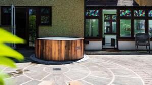 Un bain à remous en bois, installé sur une terrasse circulaire en pierre, d’où l’on peut admirer un bâtiment aux fenêtres ornées de vitraux.