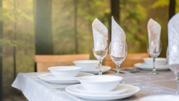 Une table dressée avec de l’assiette blanche, des bols et de la verrerie transparente, sur fond naturel flou.