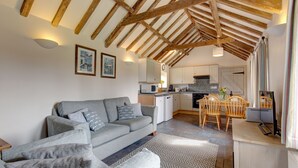 Een gezellige woonkamer met een grijze bank, houten balken in het plafond, een keuken met witte kasten en een eettafel met stoelen.