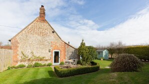 A brick house with a stone chimney, a small door, and a garden with a hedge and a tree.