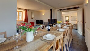 Un comedor con una larga mesa de madera dispuesta para seis personas, rodeada de sillas de madera. Hay una jarrón blanco con flores sobre la mesa, y se ve un televisor al fondo.