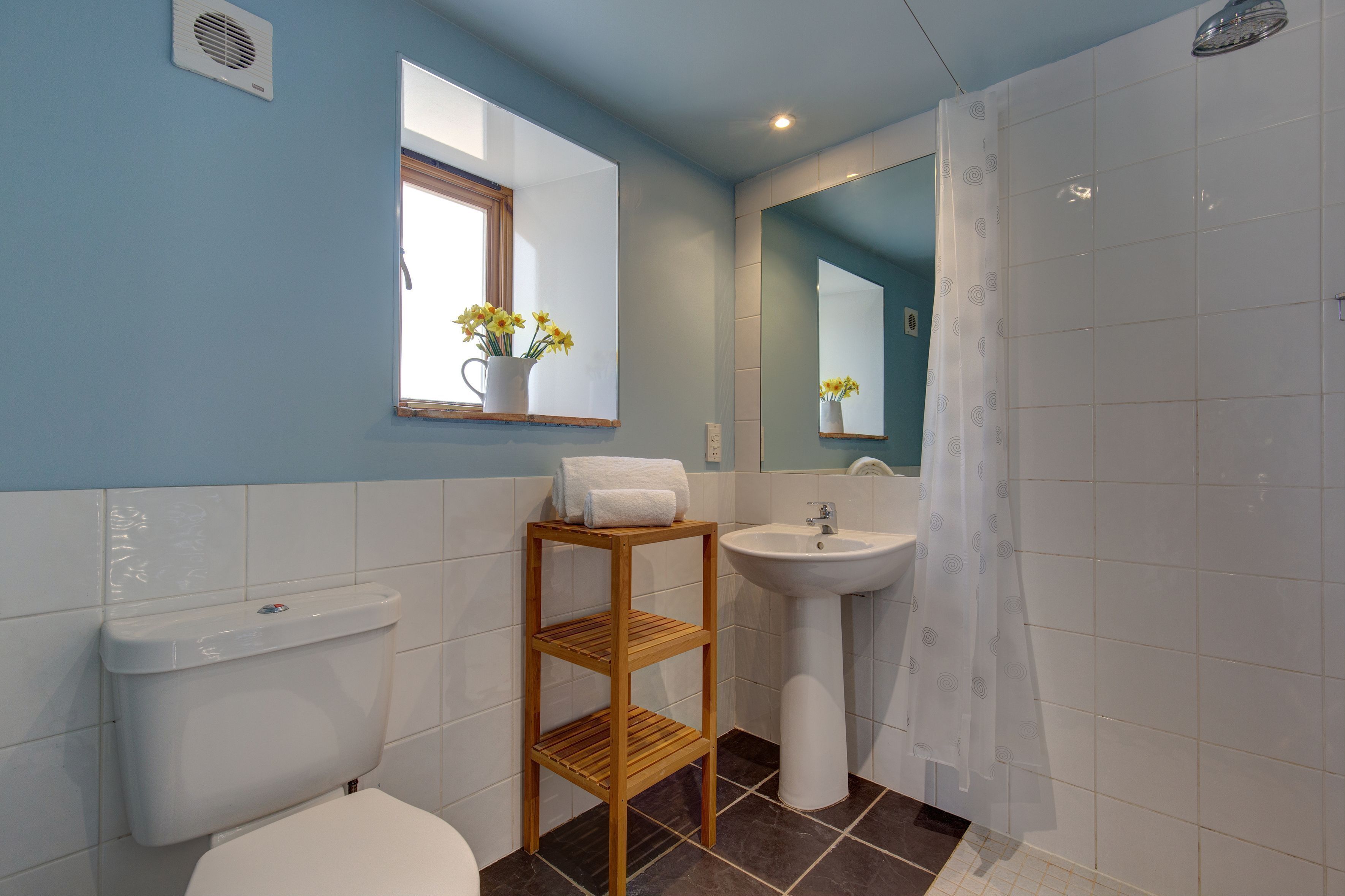 A bathroom with a toilet, sink, and shower. There is a wooden shelf with a vase of flowers and a towel on it.