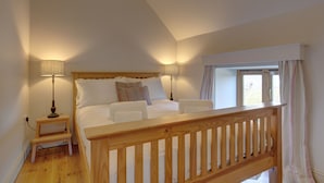 A bedroom with a wooden bed frame, two bedside lamps, a window with curtains, and a small wooden stool. - Workshop - sleeps 8 guests  in 4 bedrooms (Holt)