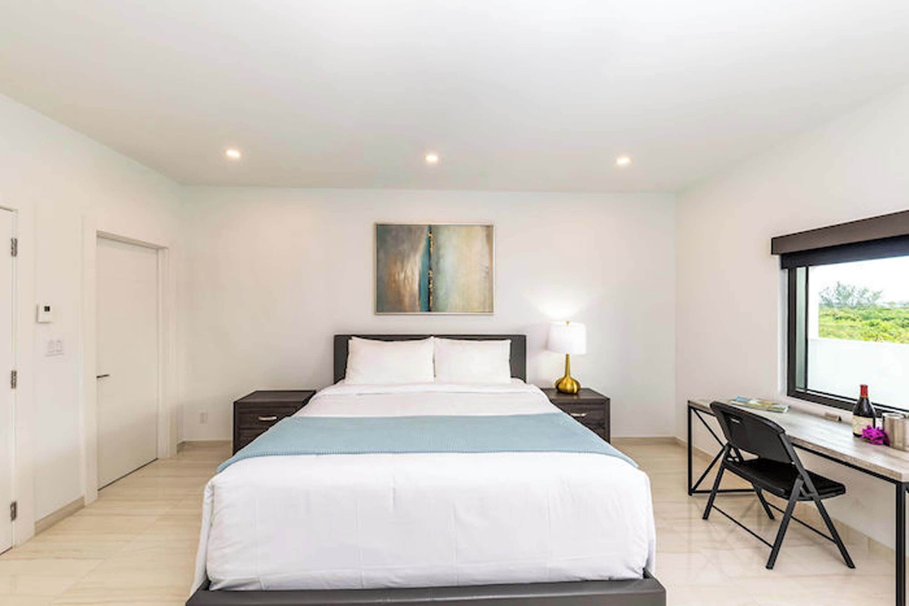 A modern hotel room with a large bed, a desk with a chair, a nightstand with a lamp, and a window with a view.