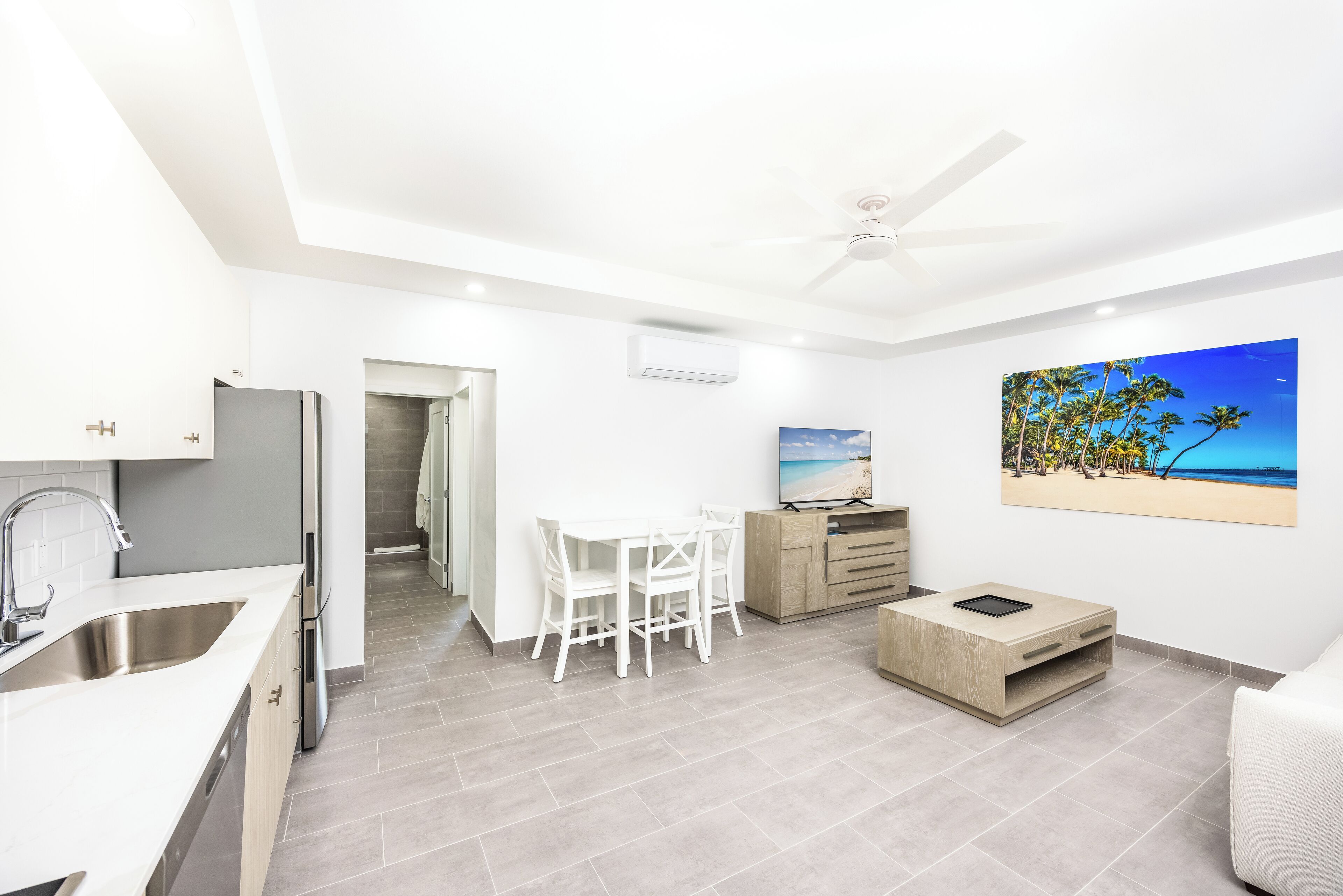 A modern kitchen with a built-in dishwasher, a dining area with a white table and chairs, a ceiling fan, and a beach-themed painting on the wall.
