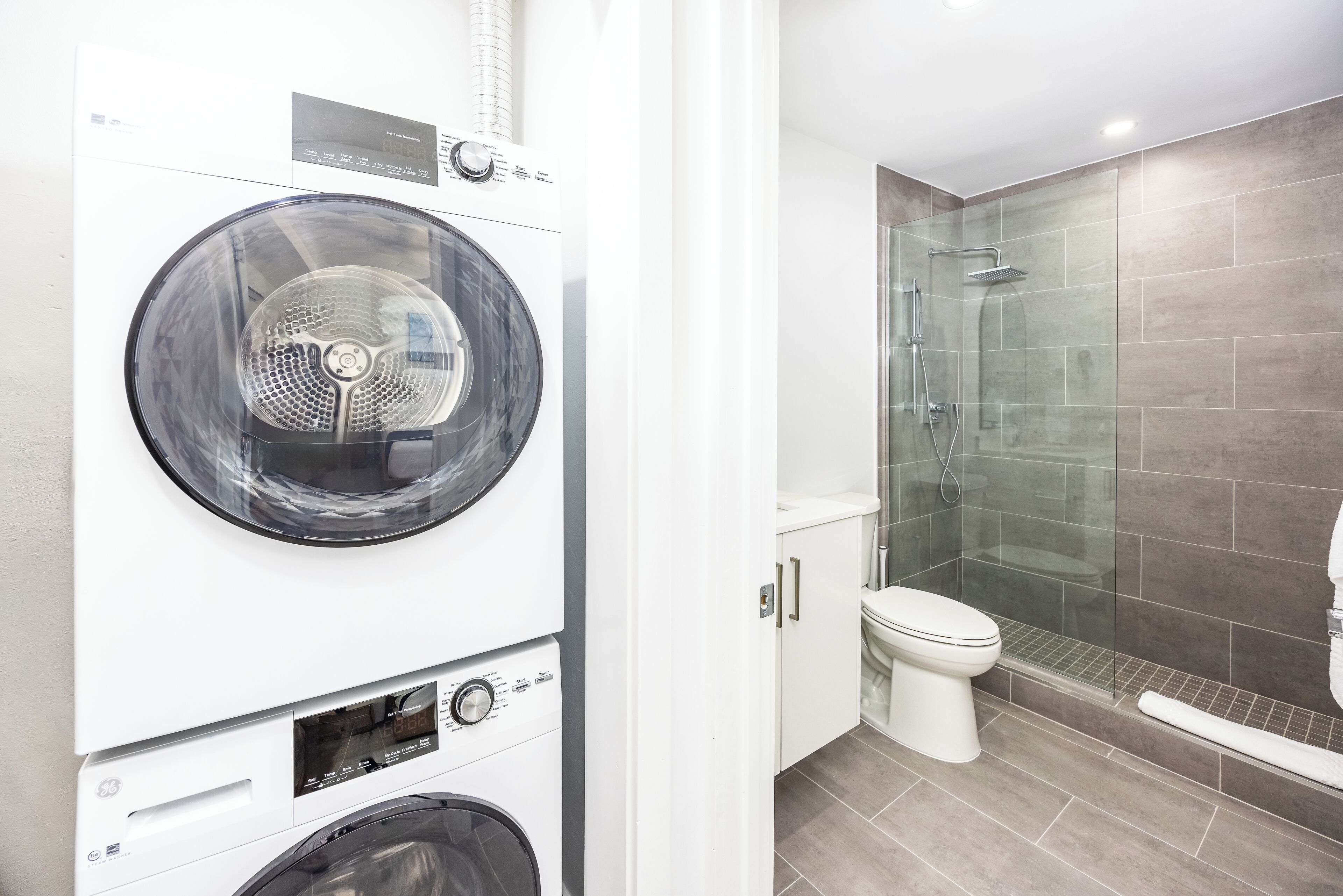 Un espace buanderie moderne équipé d’un lave-linge et d’un sèche-linge à chargement frontal, et d’une douche en verre située à côté des toilettes.