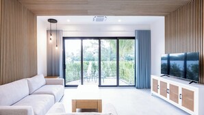 A modern living room with a sofa, a coffee table, a TV on a cabinet, and a sliding glass door leading to a balcony with outdoor furniture. - Seascape Grace Bay Hotel & Townhome Villas (Providenciales)