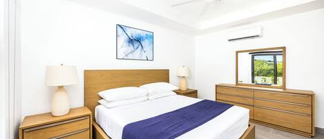 Et moderne soveværelse med en træseng, hvide sengetøj, et blåt tæppe, en loftsvifte, et natbord med lampe, en kommode med skuffer, et spejl og et vindue med udsigt.