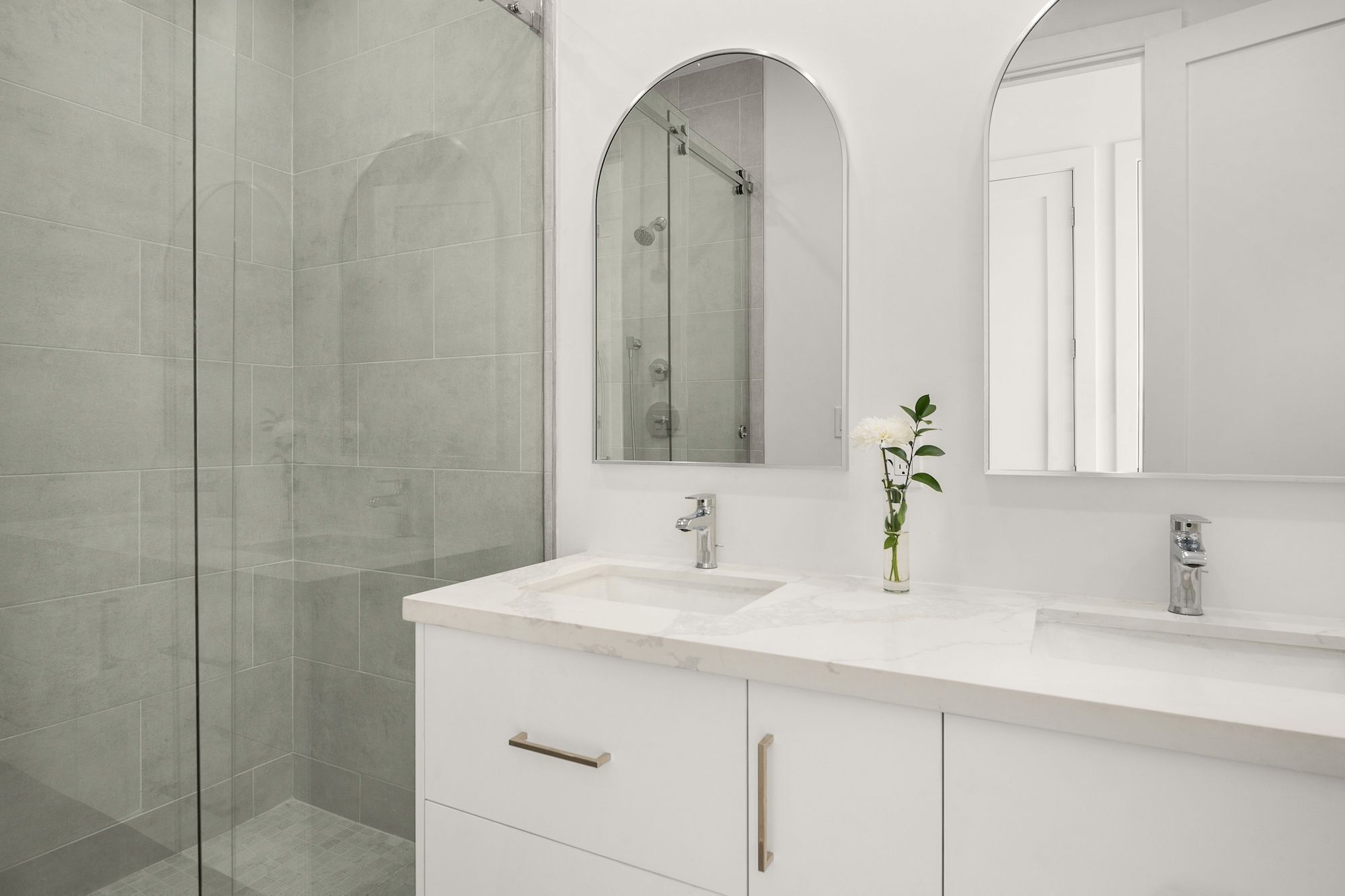 A modern bathroom with a glass shower enclosure, a white vanity with a marble countertop, and a single white flower in a vase.