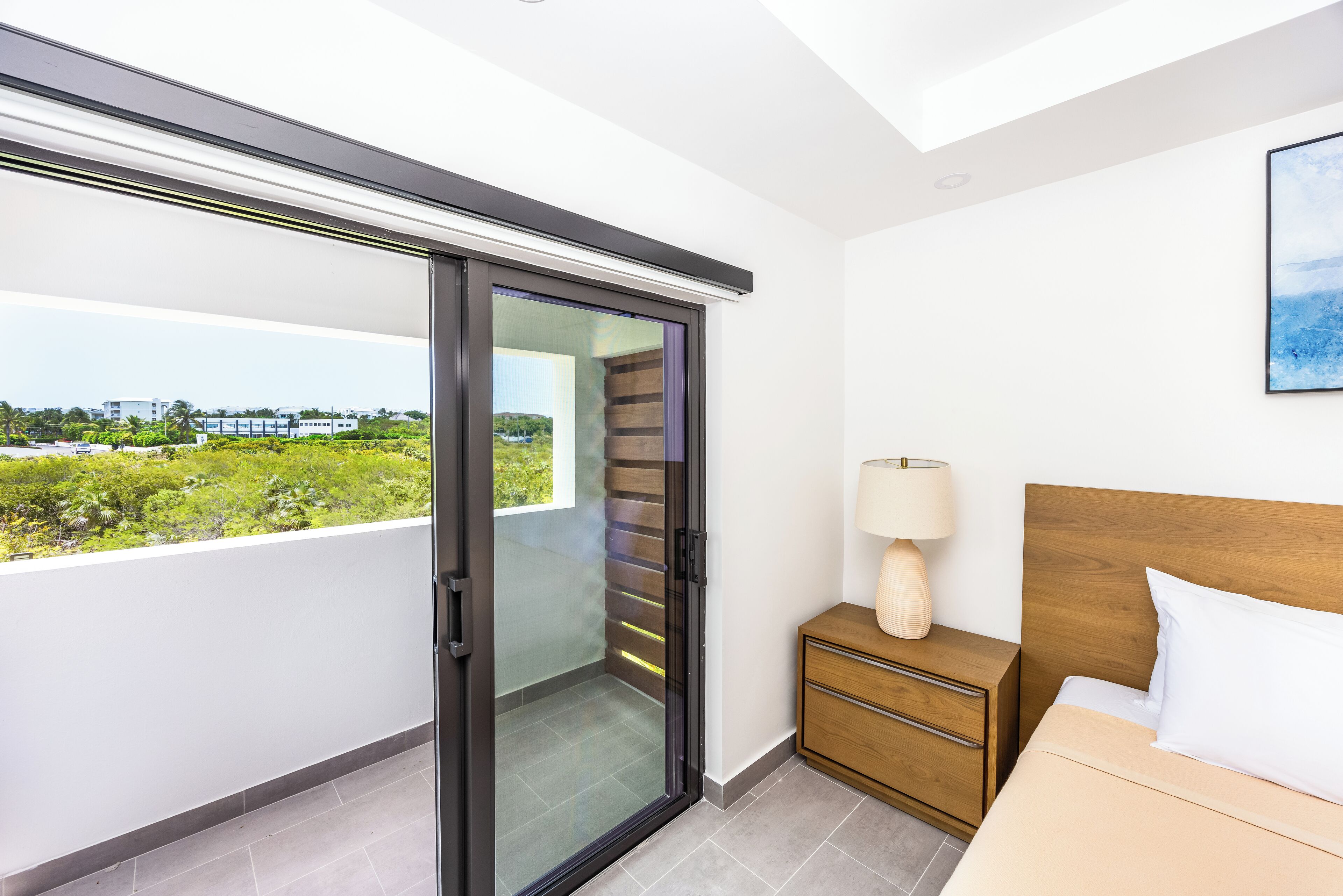 Ein Schlafzimmer mit Bett, Nachttisch und Blick durch eine Glastür auf einen Balkon mit Stadtansicht.