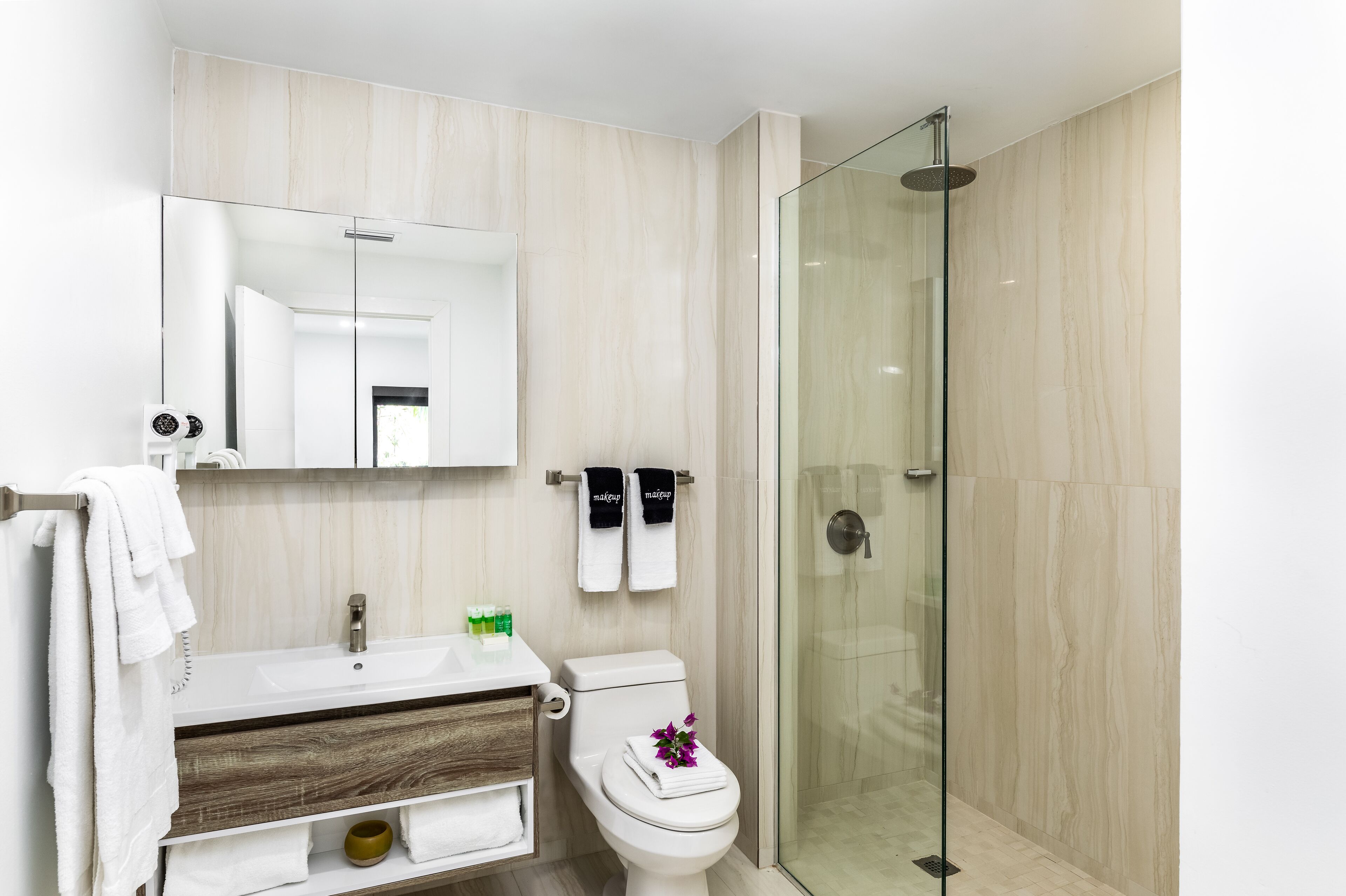 Ein modernes Badezimmer mit einer gläsernen Duschkabine, einem weißen Waschbecken mit Holzunterschrank und einer Toilette, auf der ein Handtuch und eine Blume liegen.