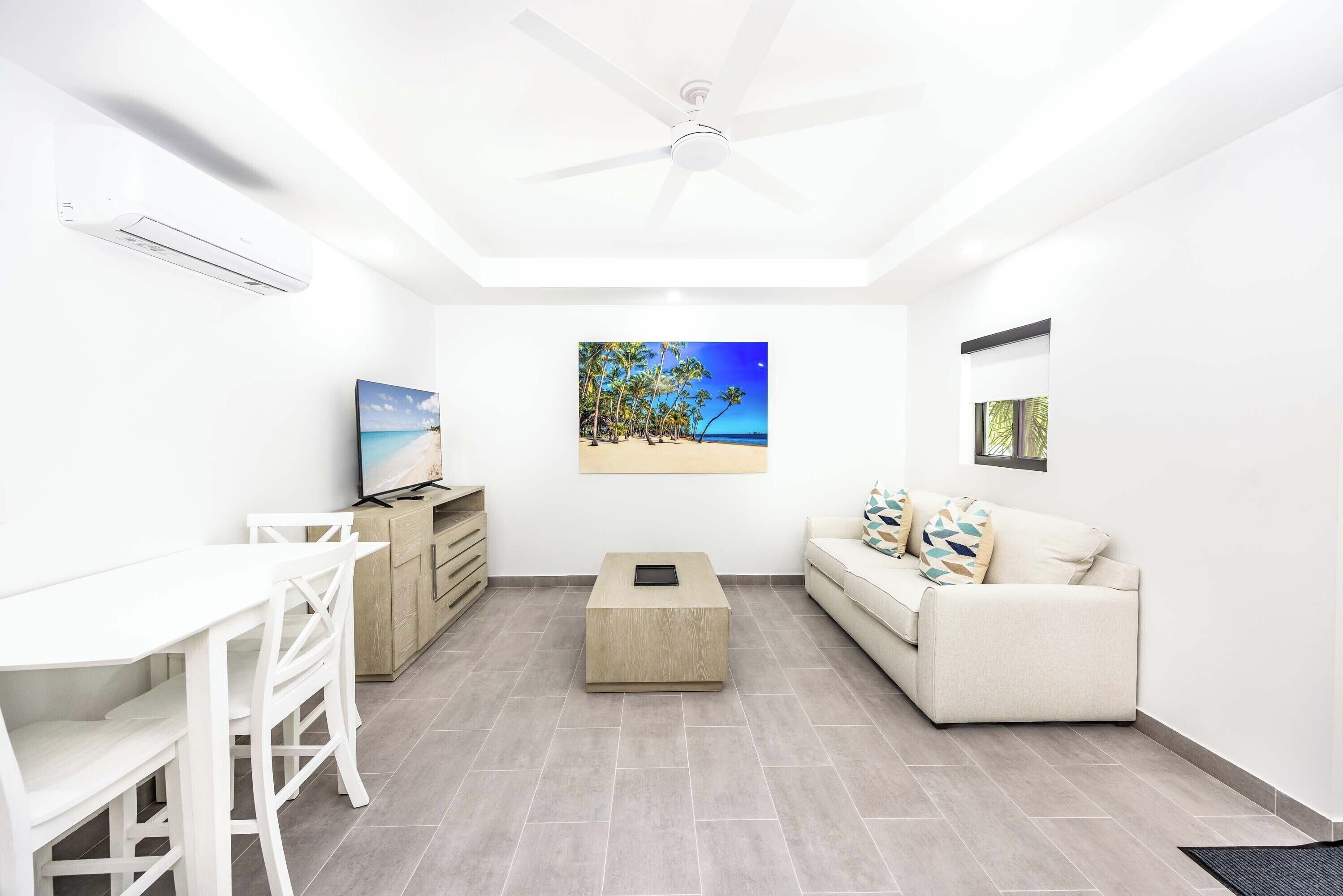 A modern living room with a white ceiling fan, a flat-screen TV, a wooden coffee table, a white sofa, and a dining area with white chairs.