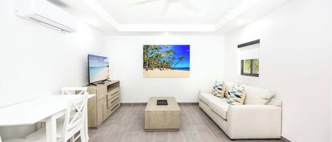 A modern living room with a white ceiling fan, a flat-screen TV, a wooden coffee table, a white sofa, and a dining area with white chairs.