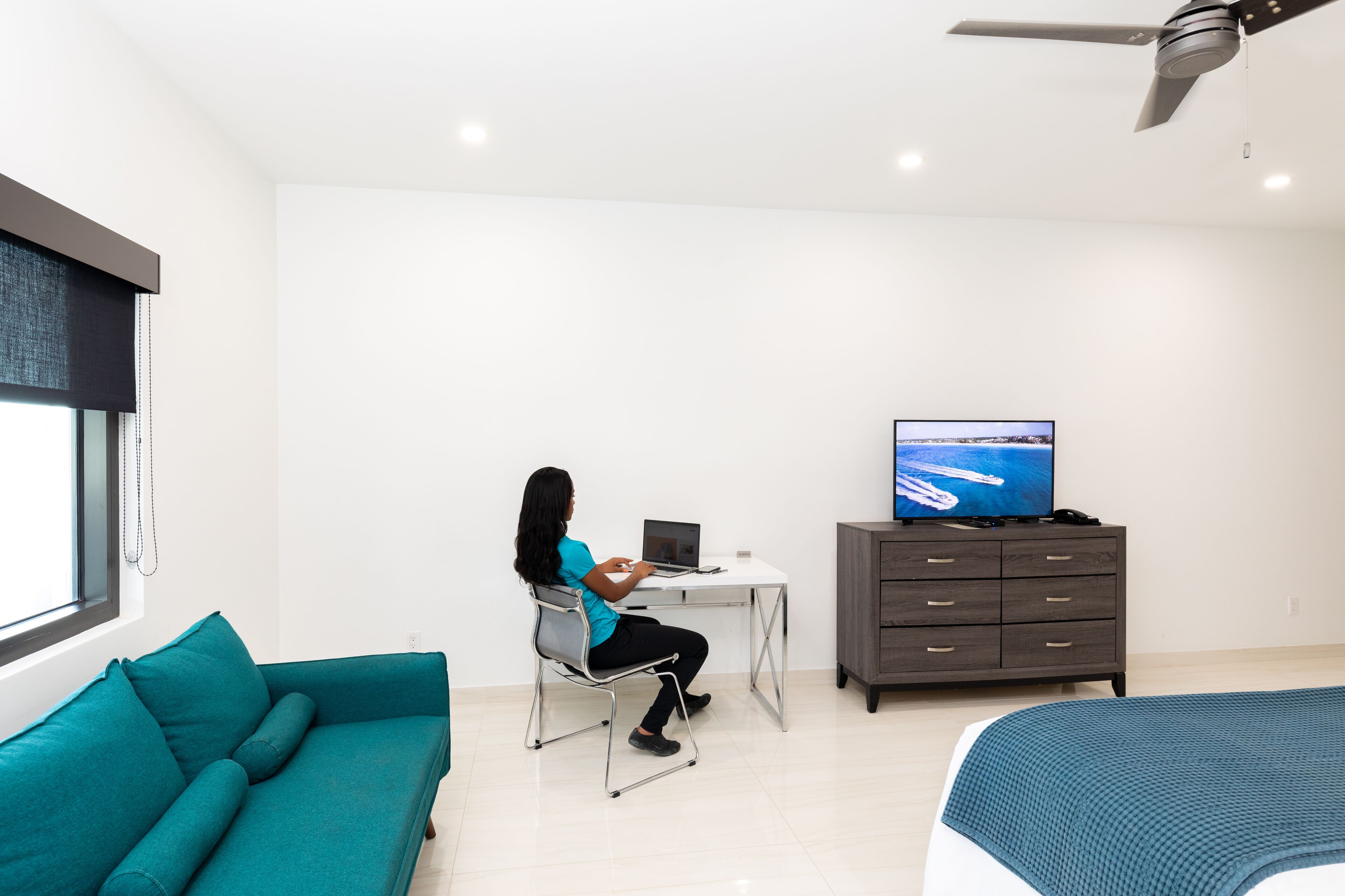 Un dormitorio moderno con un sofá azul aguamarina, un escritorio con una computadora portátil, un televisor y una cama con una manta azul.