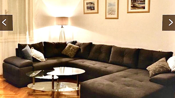 Ein Wohnzimmer mit einem Ecksofa, einem Couchtisch und einer Stehlampe. An der Wand hängen drei gerahmte Bilder.