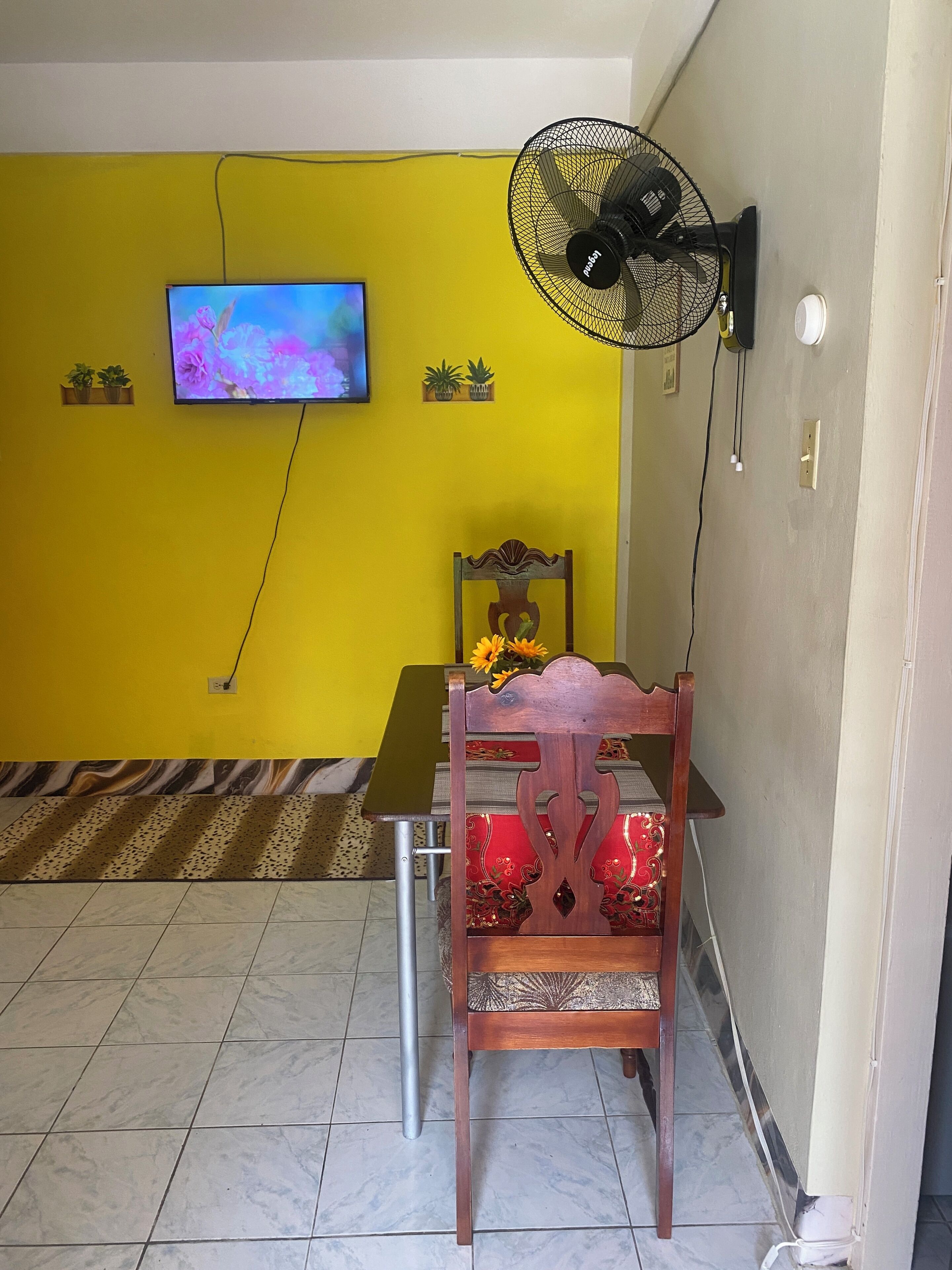 Ein Zimmer mit gelber Wand, einem daran befestigten Fernseher, einem Standventilator, einem Holzstuhl mit Kissen und einem gemusterten Teppich auf dem Boden.