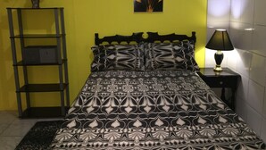 Una cama con colcha estampada, una mesita de noche con lámpara, un cuadro en la pared y una estantería con un jarrón de flores.
