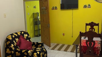A room with a sofa covered in floral upholstery, a dining table with a red tablecloth, a wooden chair, a television mounted on the wall, and a fan.