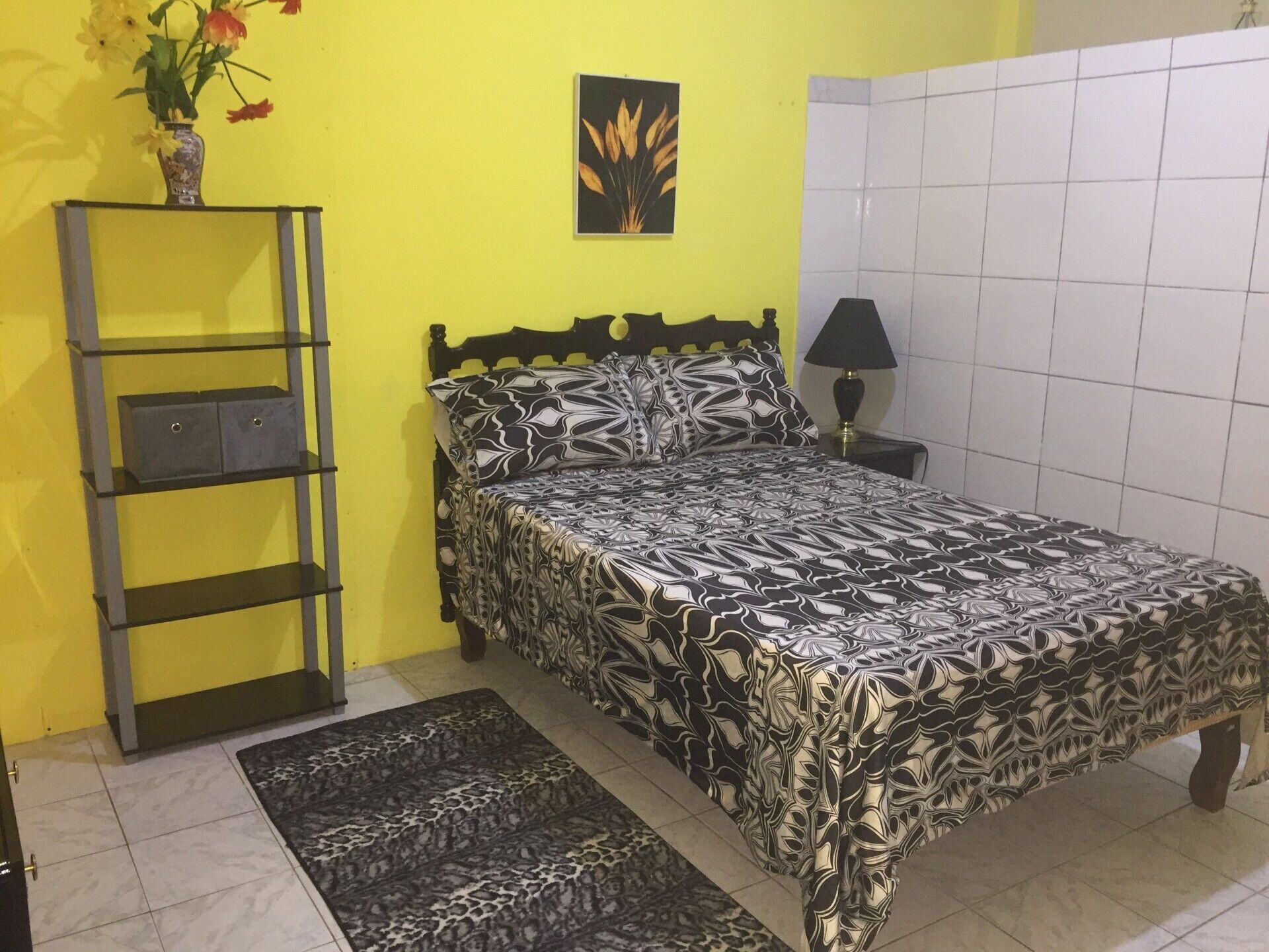 Una cama con edredón estampado, una mesita de noche con lámpara, una estantería con cajas de almacenamiento y un jarrón con flores.