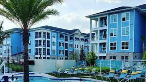 Une piscine entourée d’un bâtiment bleu doté de nombreux balcons, d’un palmier et de chaises longues.