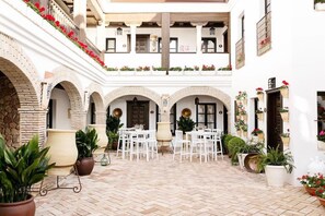 Un patio con suelo de baldosas blancas, sillas blancas y plantas en macetas.