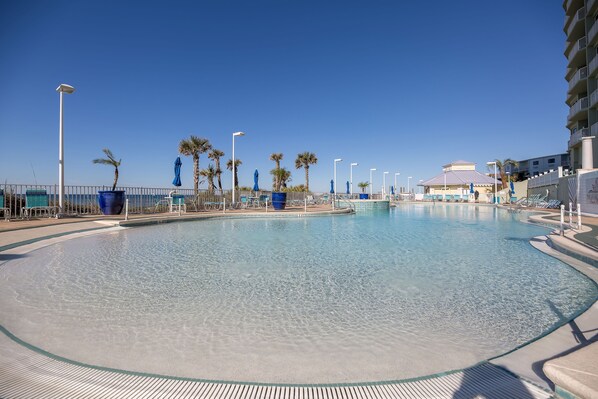 Une grande piscine aux eaux cristallines, bordée d’une terrasse carrelée et entourée de palmiers et de parasols bleus.