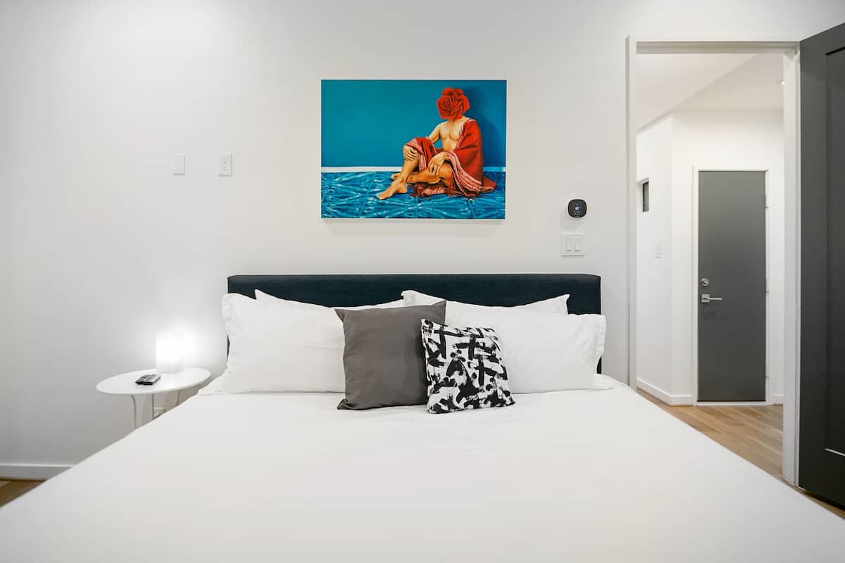 A hotel room with a bed, white bedding, a blue wall, and a painting of a person sitting on the floor.