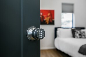 Una puerta moderna con cerradura electrónica, una cama blanca con un cojín estampado y un cuadro abstracto rojo en la pared.