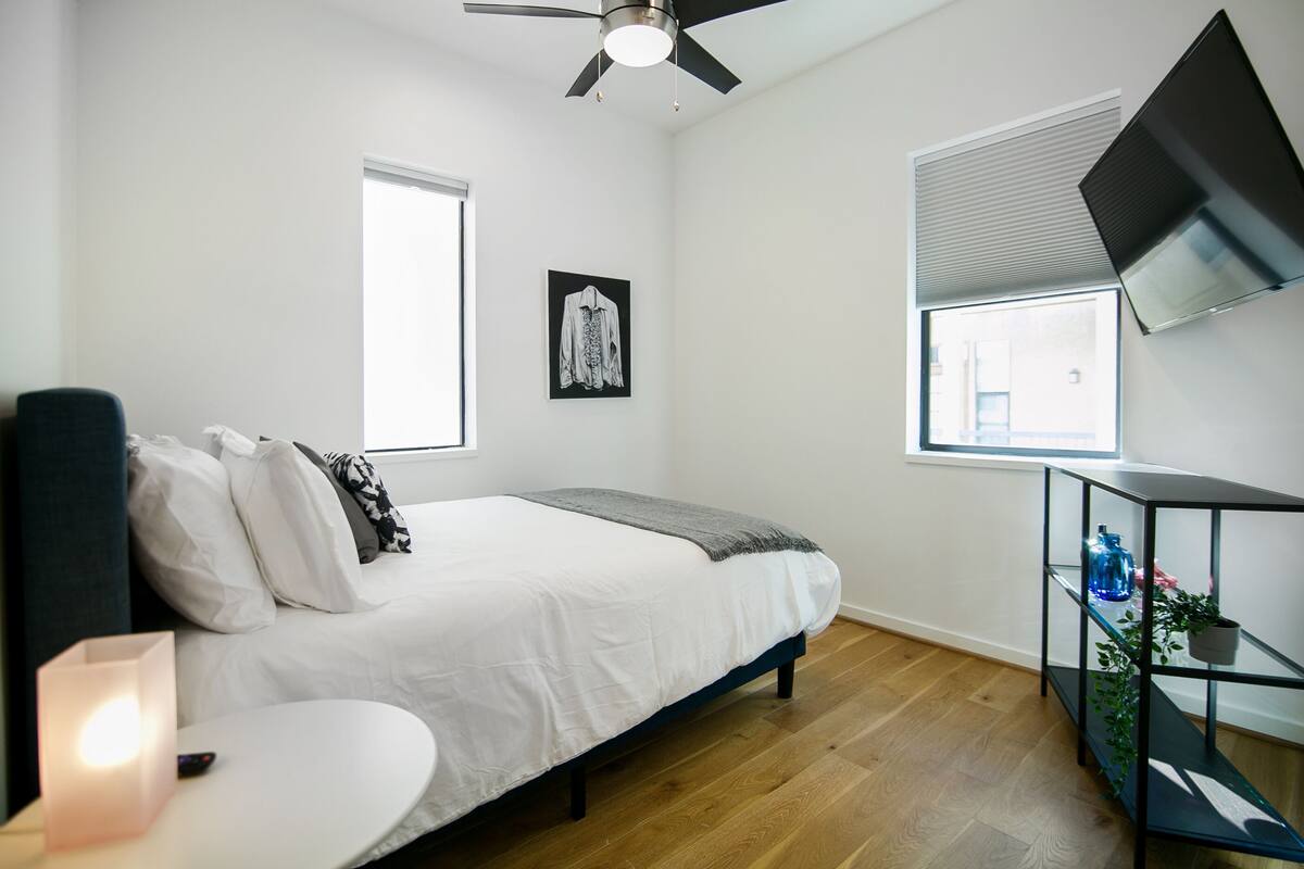A bedroom with a bed, a bedside table with a lamp, a TV mounted on the wall, and a window with blinds.