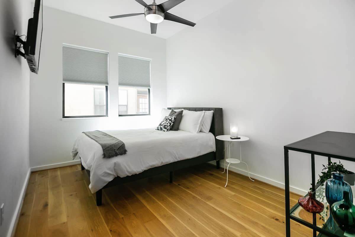 A bedroom with a bed, a ceiling fan, a small table with a lamp, and a shelf with decorative items.